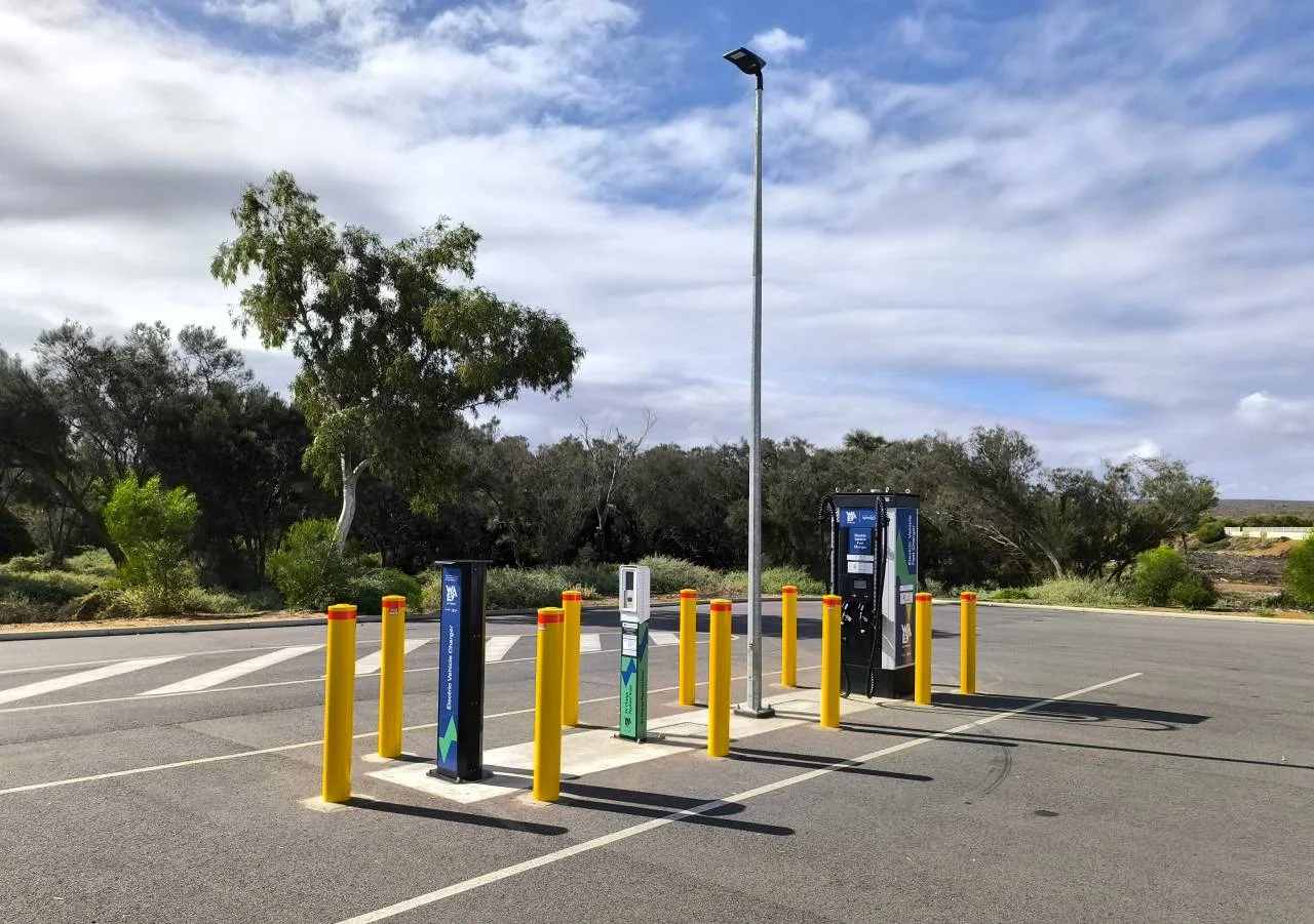 An electric vehicle charging station with multiple chargers and yellow bollards in an empty parking lot, surrounded by trees and under a partly cloudy sky.