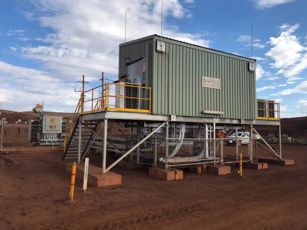 A metal industrial control room stands elevated on steel stilts with stairs and yellow railings, surrounded by red dirt and utility equipment under a blue sky with scattered clouds.
