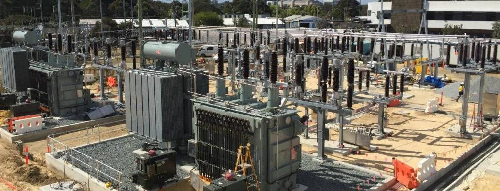 A large electrical substation with multiple transformers, circuit breakers, and power lines, surrounded by safety barriers and construction equipment, set in an urban area with buildings and trees in the background.