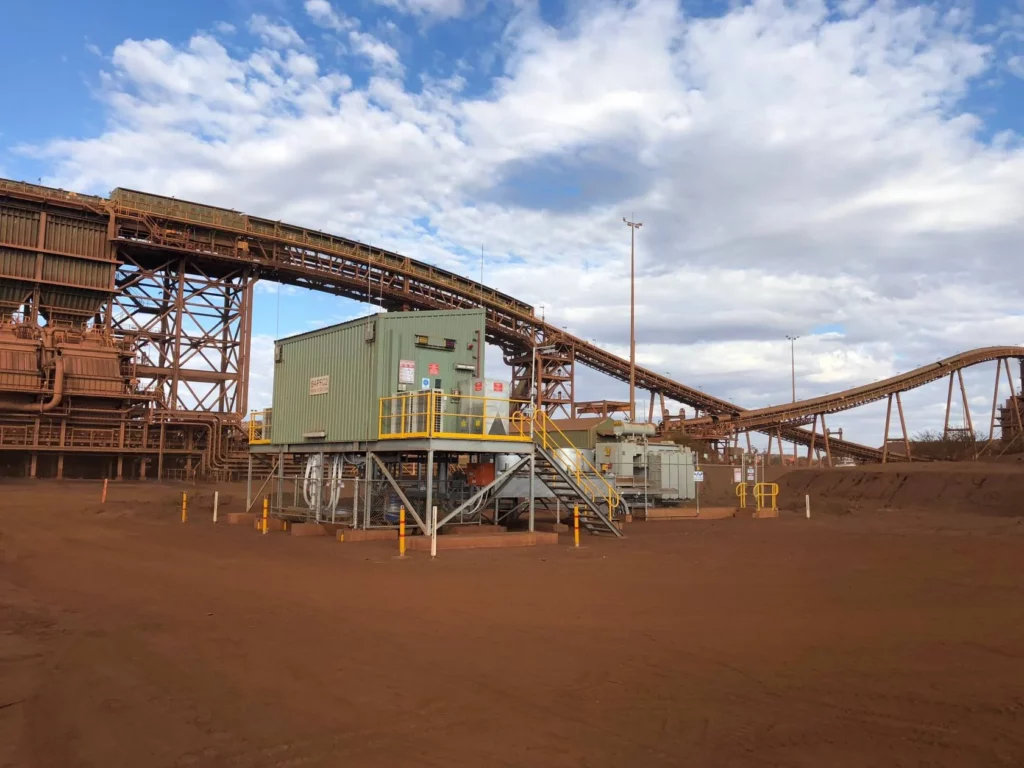 A mining site with a large industrial conveyor system, steel structures, and green control cabins on red dirt under a partly cloudy sky. Yellow railings and poles mark pathways around the equipment.