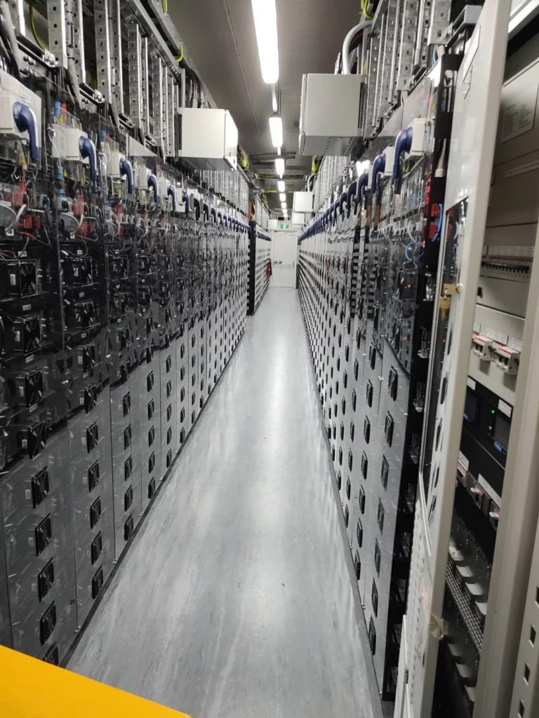 A brightly lit aisle in a data center, lined on both sides with tall racks of servers and network equipment, with cables and hardware components visible.