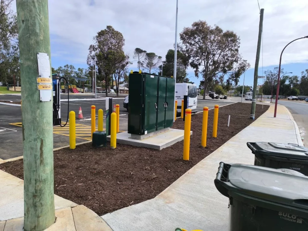 A sidewalk with green utility boxes surrounded by yellow bollards, next to a parking lot. Trash bins are on the pavement, and trees and parked vehicles are visible in the background under a cloudy sky.