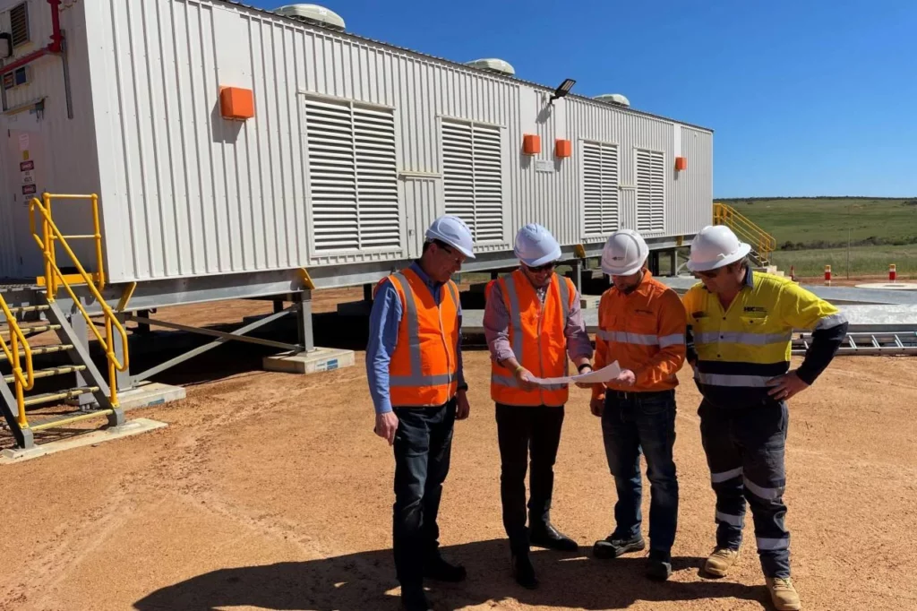 Four construction workers wearing hard hats and high-visibility vests stand on a dirt site, examining documents in front of a large portable building under a clear blue sky.