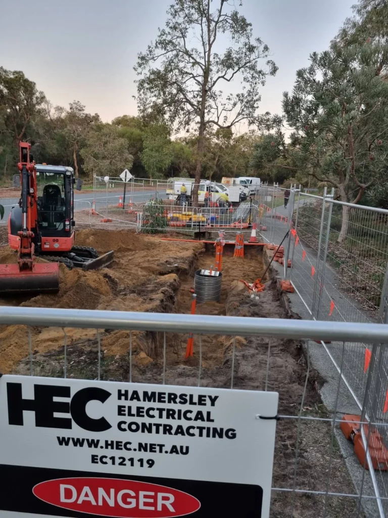 A fenced construction site with a red excavator, dug trenches, orange safety barriers, and a "Danger" sign for Hamersley Electrical Contracting. Trees and parked vehicles are visible in the background.